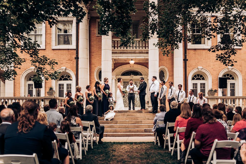 A wedding ceremony takes place on the steps of a historic peach-colored mansion with columns.