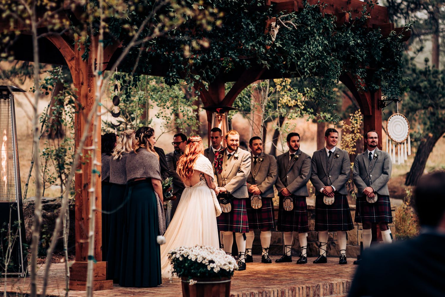 Wedding party stands on a wooden platform during outdoor ceremony with groomsmen wearing traditional kilts.