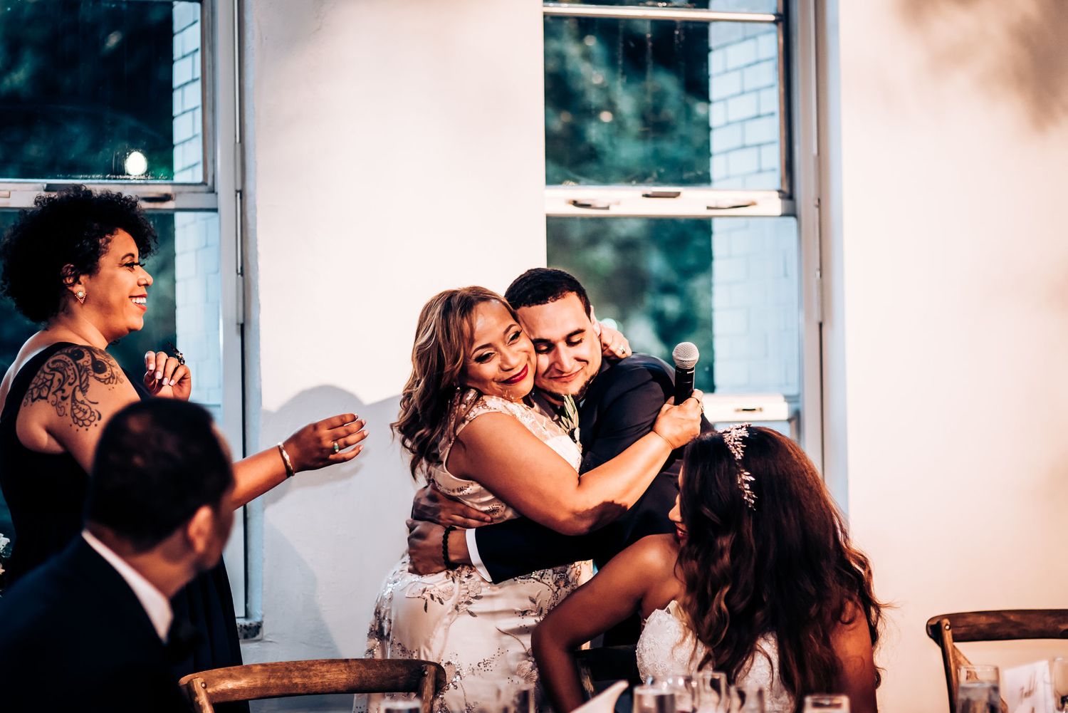 Wedding reception guests share joyful embraces during an intimate celebration by window light.
