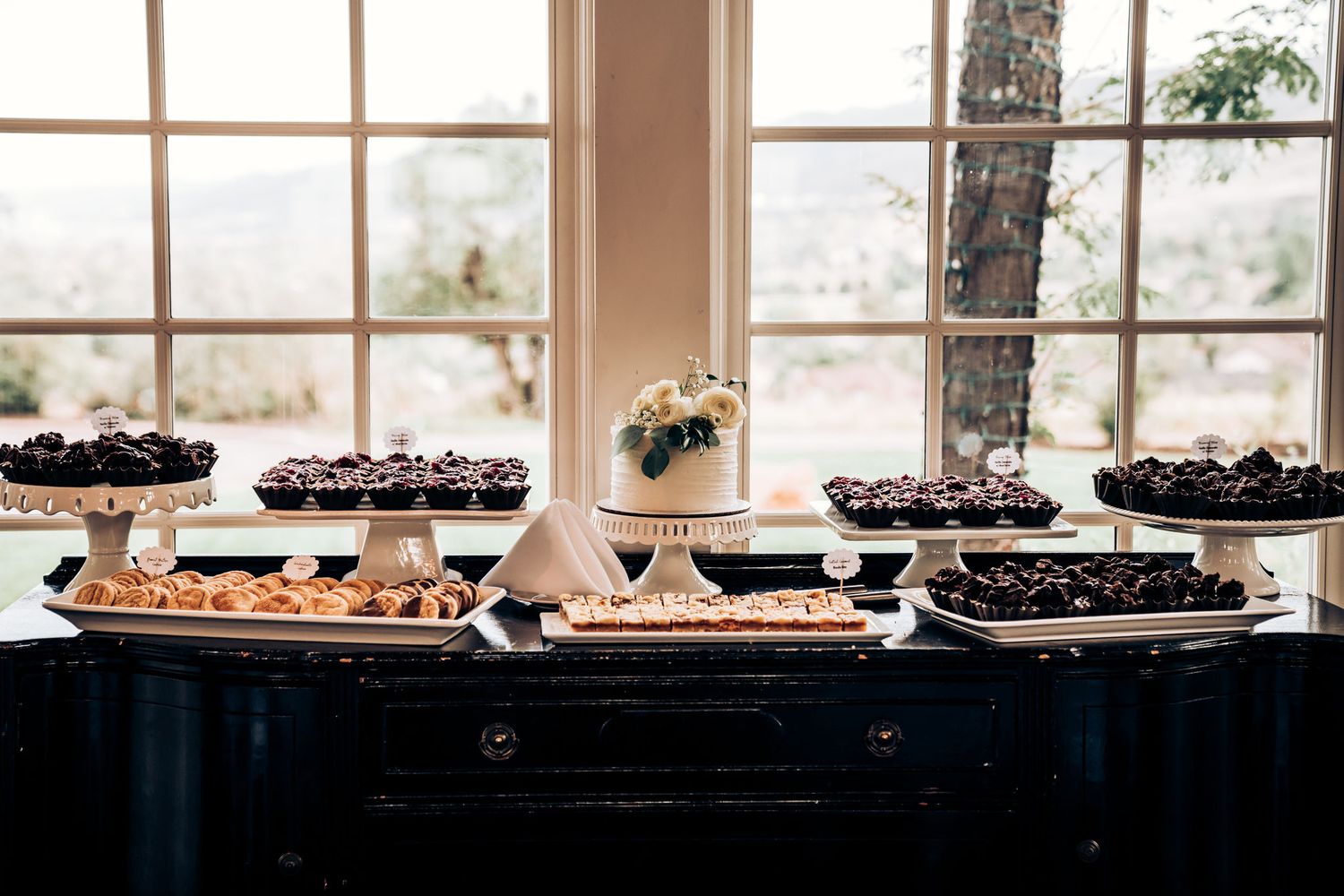 Elegant dessert table with various treats displayed near large windows.