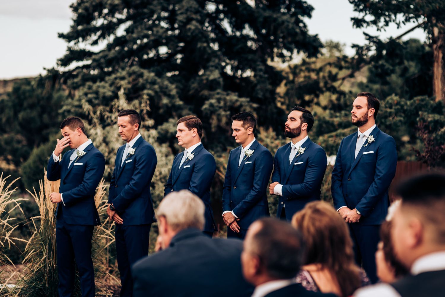 Groomsmen in navy suits standing in line during outdoor wedding ceremony.