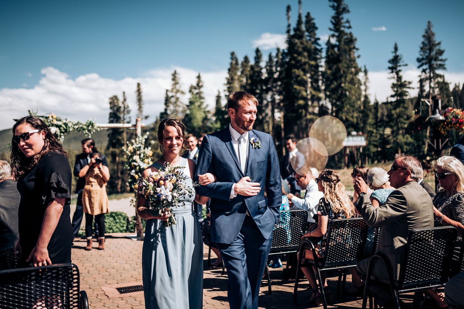 Wedding guests gather for an outdoor ceremony in front of tall pine trees on a sunny mountain day.