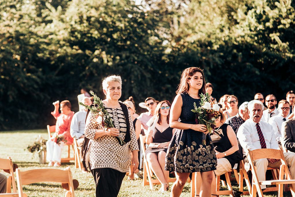 Wedding guests gather outdoors on wooden chairs during a sunny afternoon ceremony.