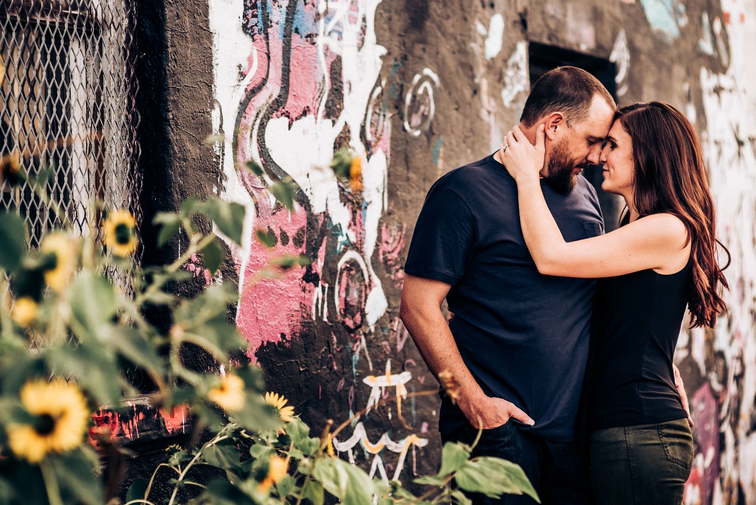 A couple embraces near a graffiti wall with yellow flowers in the foreground.