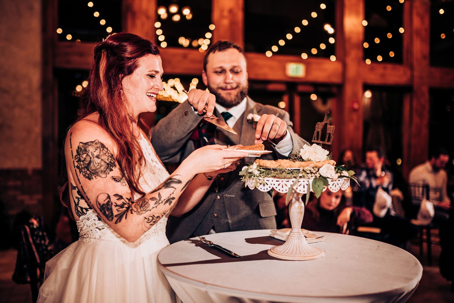 Couple shares cake-cutting moment at wedding reception in rustic venue.