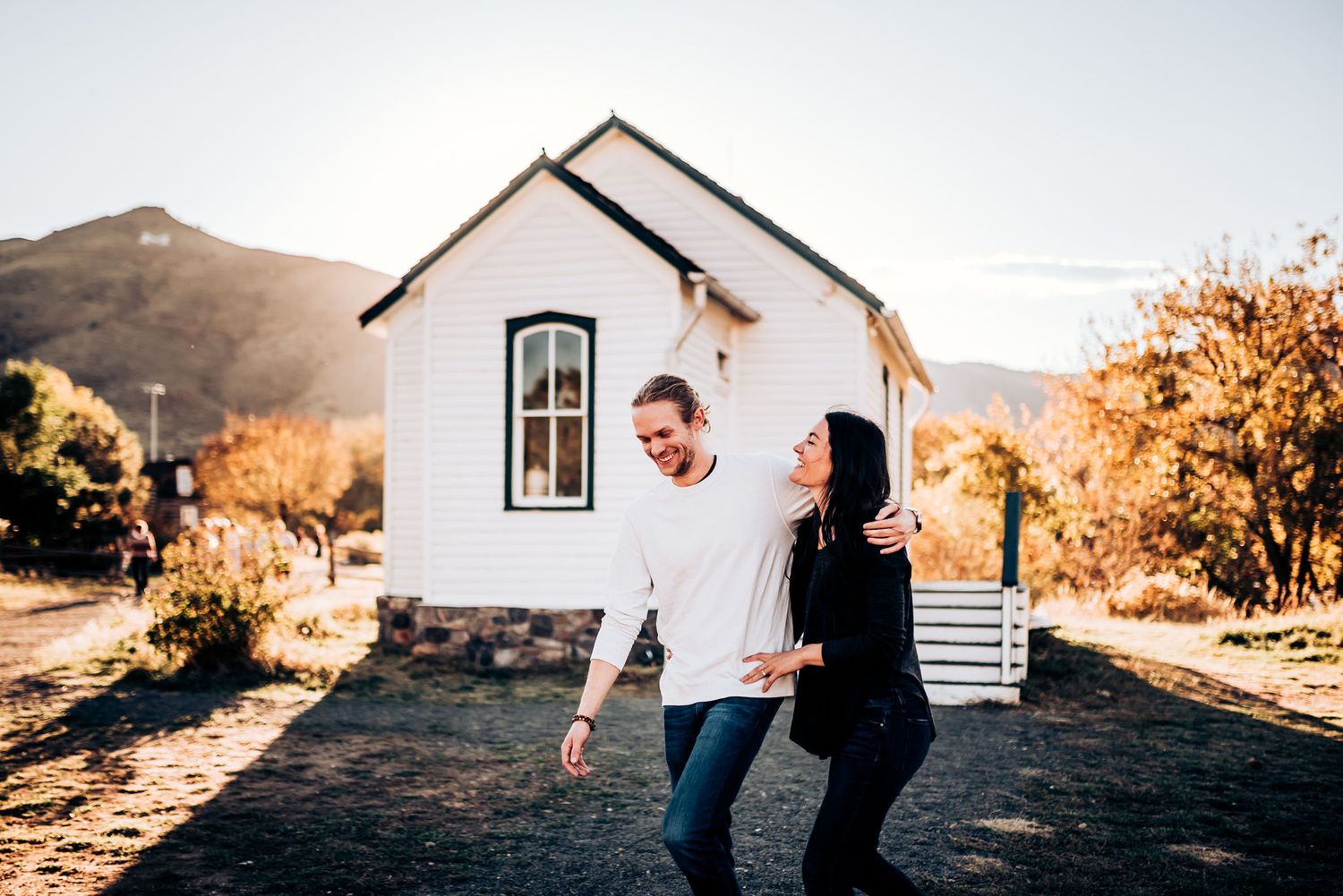 A couple playfully walks together outside a white cottage with mountains in the background.