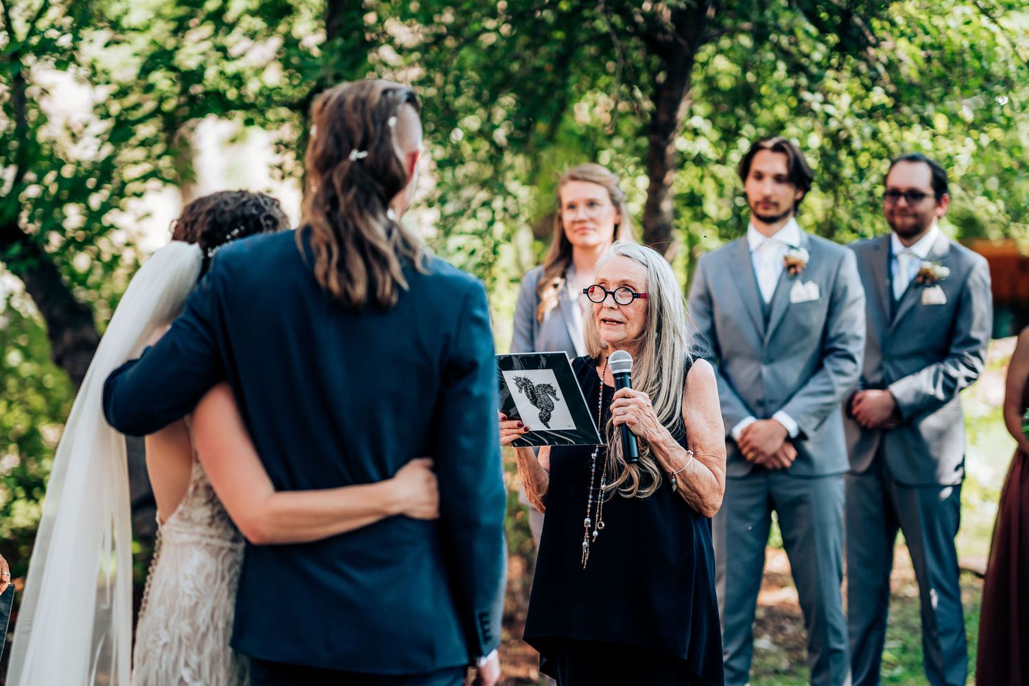 Outdoor wedding ceremony with officiant speaking to couple under tree canopy.