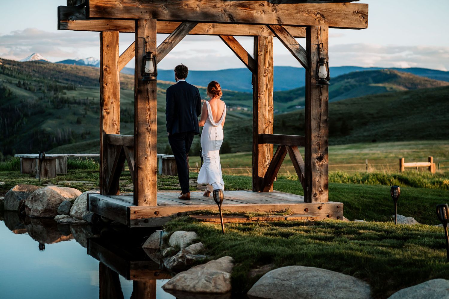 A couple stands on a wooden platform overlooking mountain vistas during sunset.