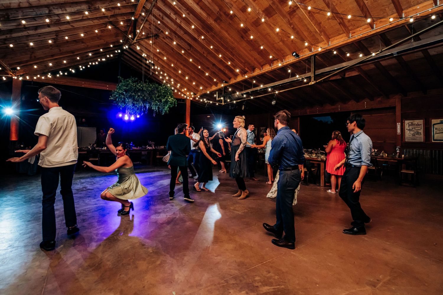 Wedding guests dancing under string lights at a rustic indoor reception venue.
