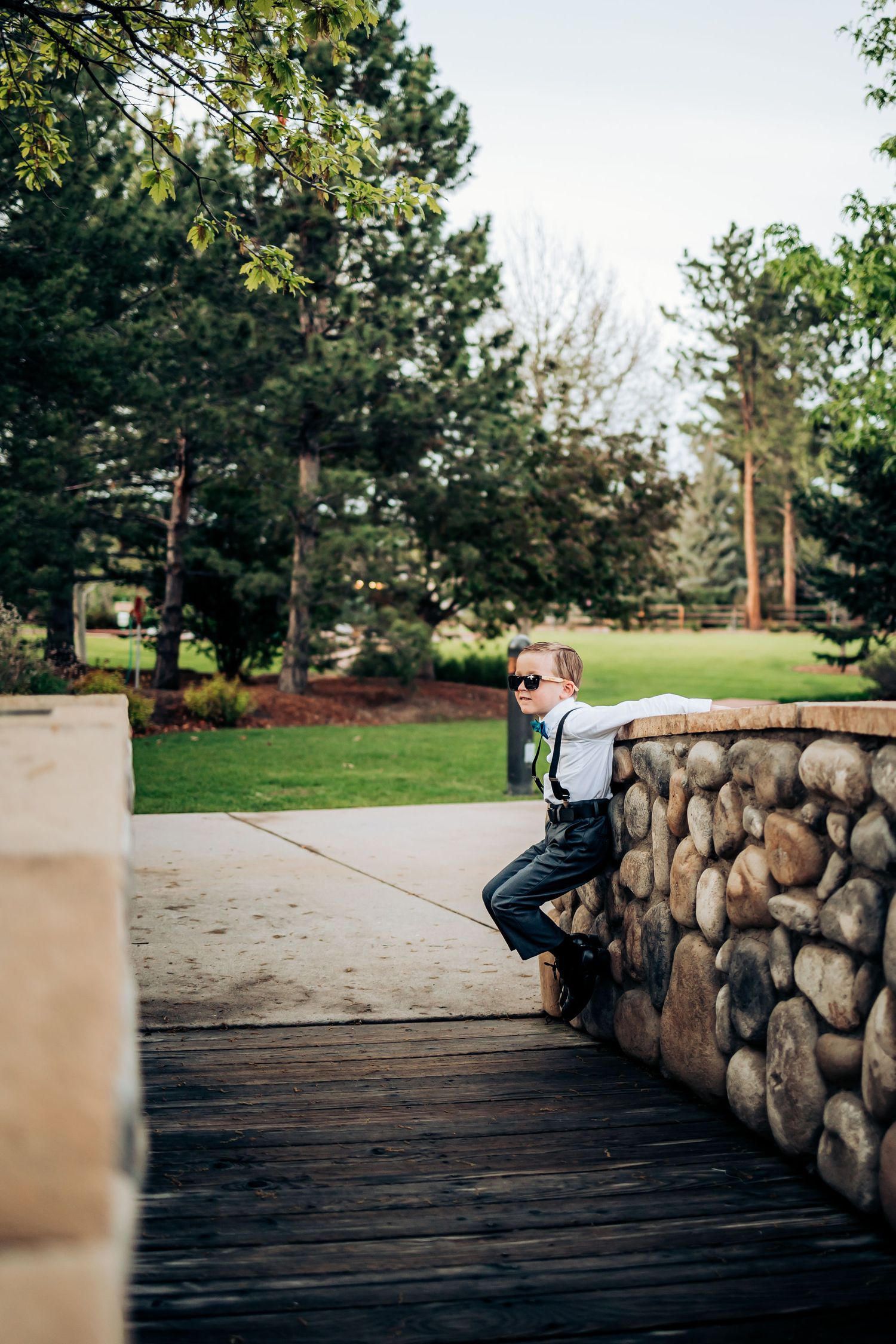 A young attendee in suspenders poses on a stone wall during a wedding.
