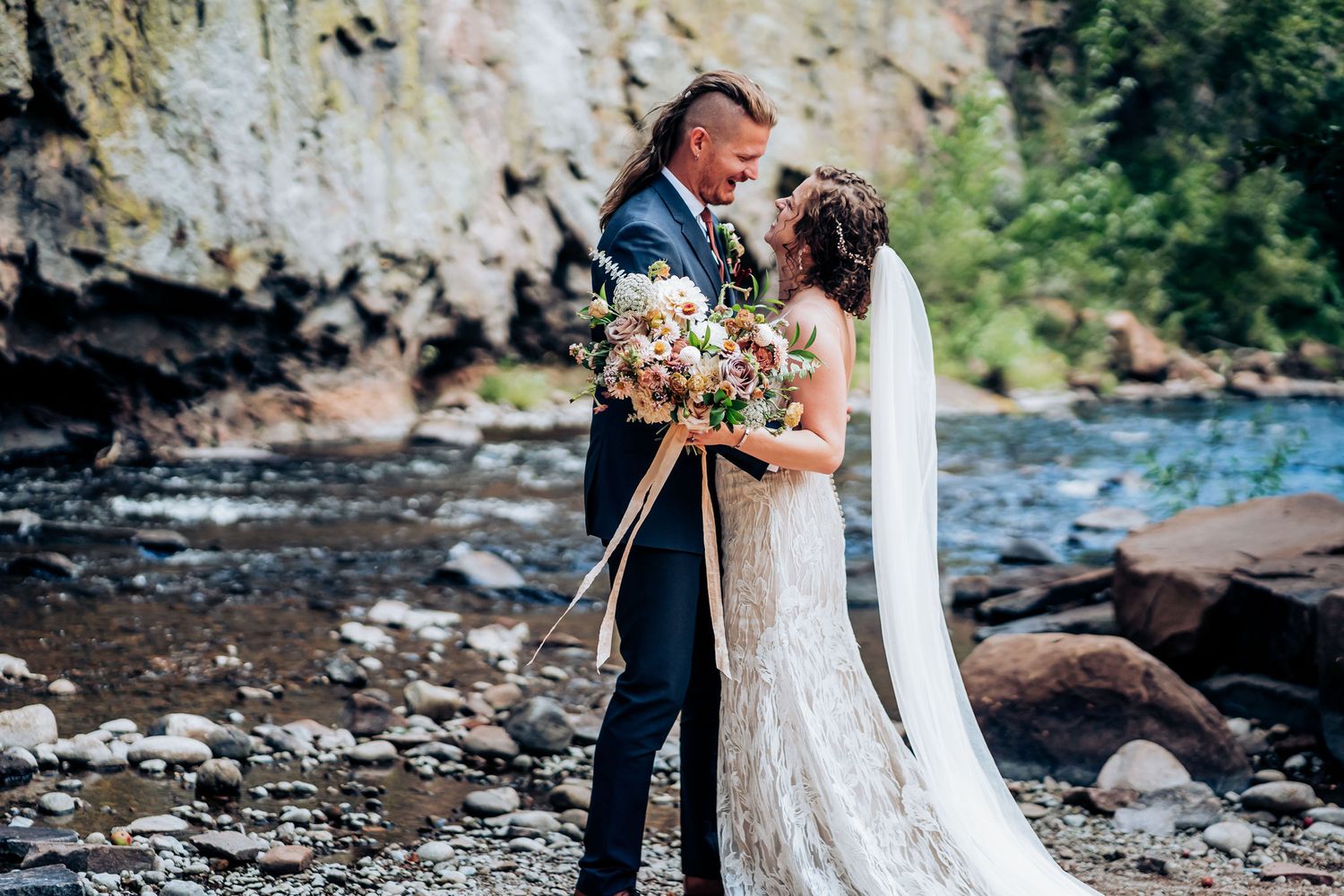 A romantic wedding couple embraces by a rocky riverbank holding a blush and cream floral bouquet.