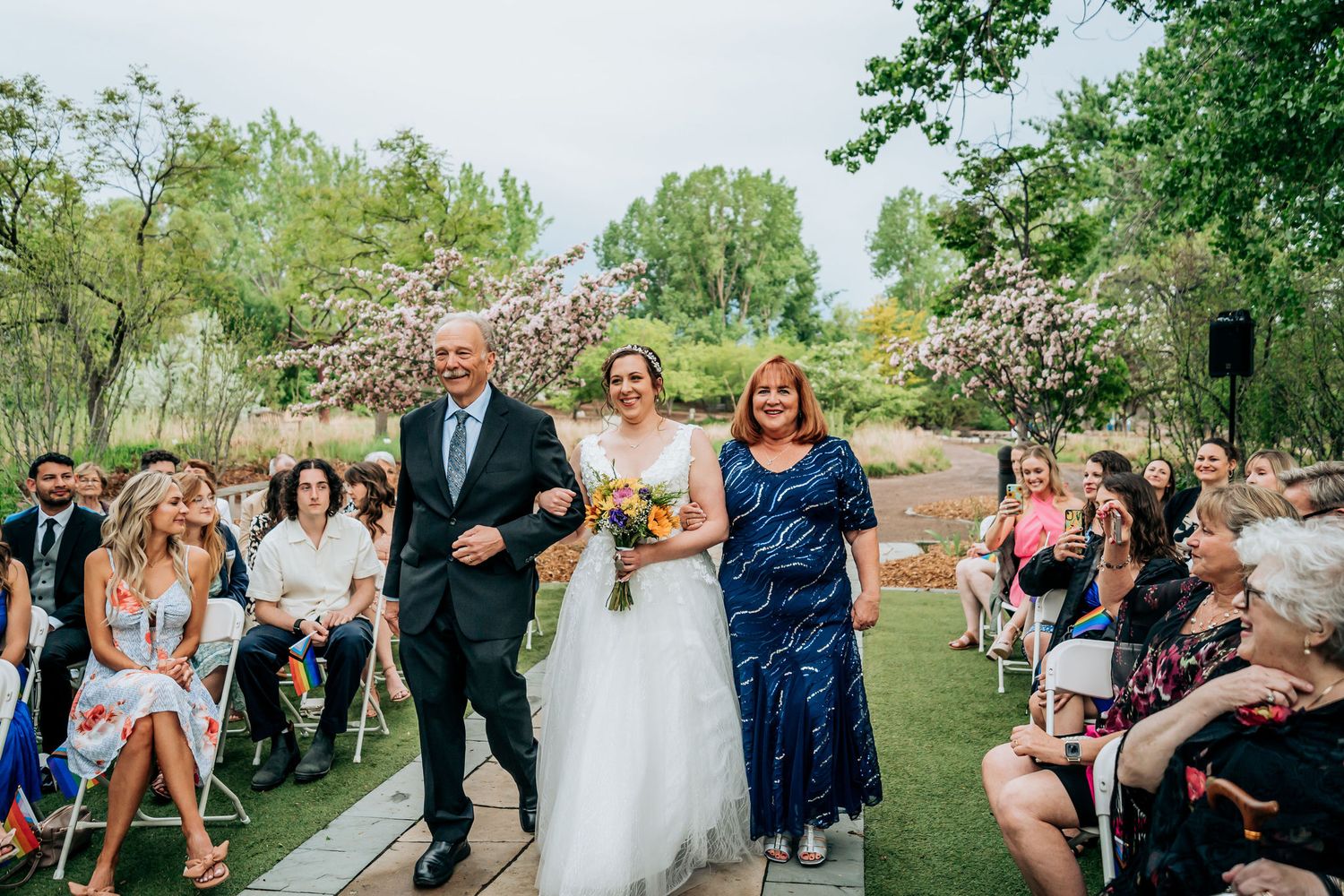 Wedding ceremony taking place outdoors on a stone path with blooming trees and seated guests in the background.