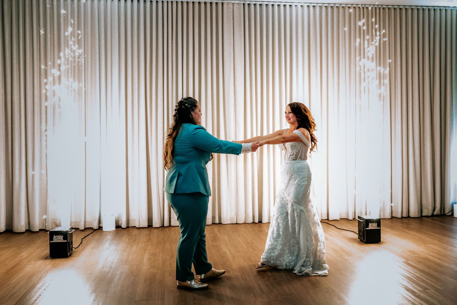 Two brides holding hands and spinning during their first dance with sparkling backdrop effects.