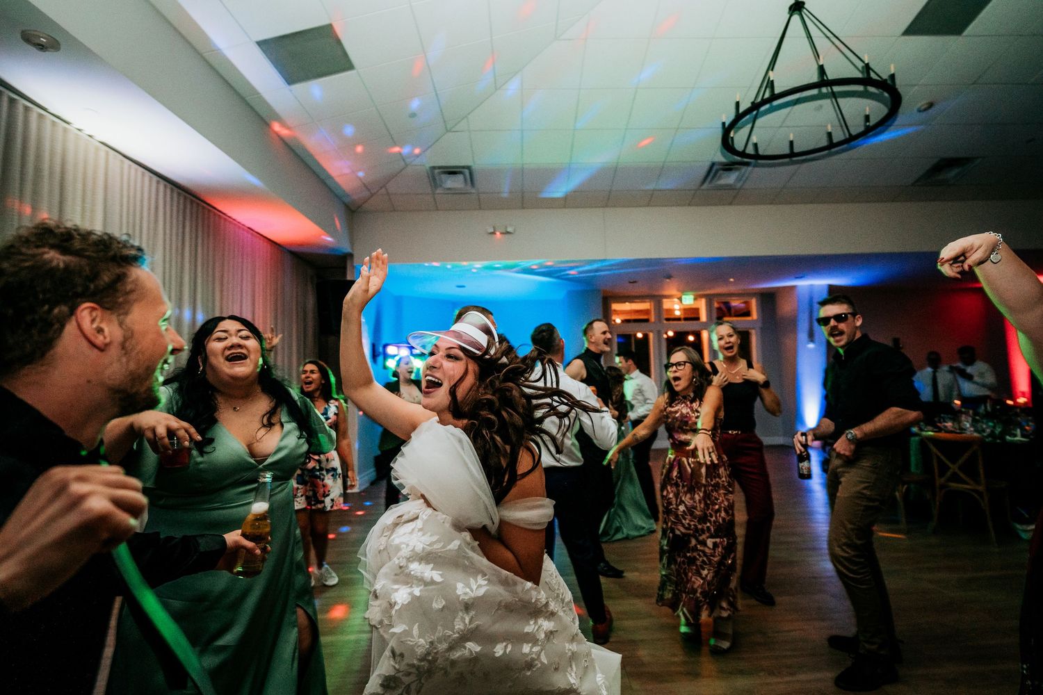 Bride and wedding guests dancing energetically at a lively indoor wedding reception with colorful lighting.