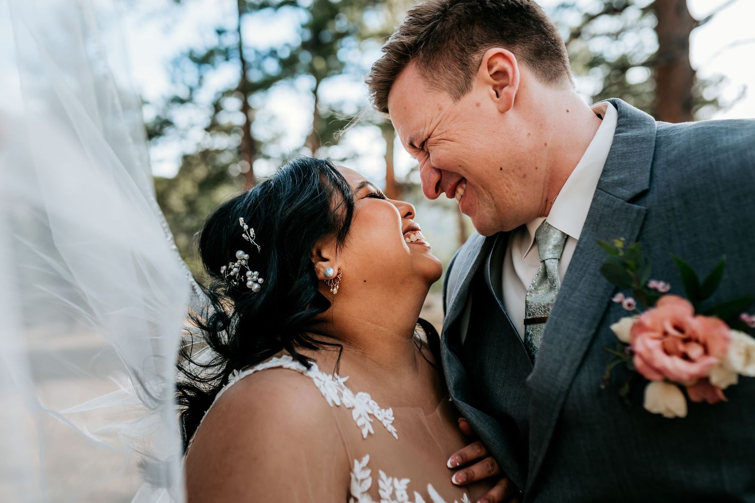 Intimate wedding moment captured between newlyweds sharing a joyful laugh under a flowing veil outdoors.