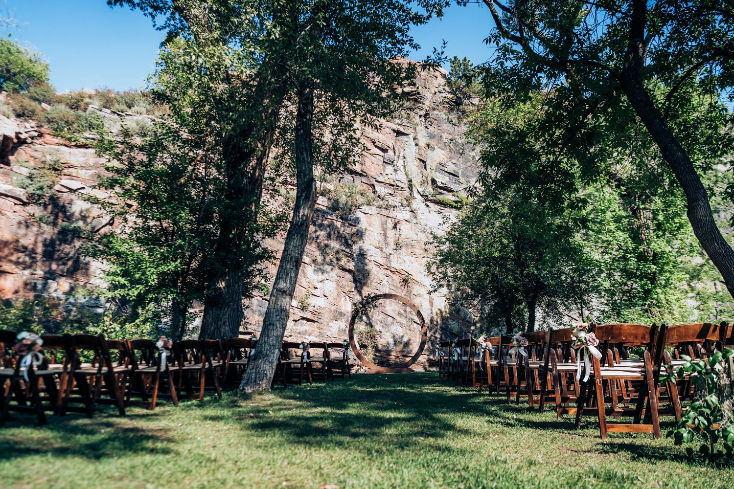 Outdoor wedding ceremony setup with wooden chairs arranged in rows facing a natural rock wall backdrop in a wooded setting.