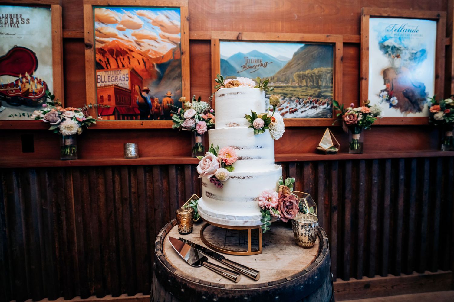 White wedding cake displayed on wooden barrel with vintage ski lodge posters in background.