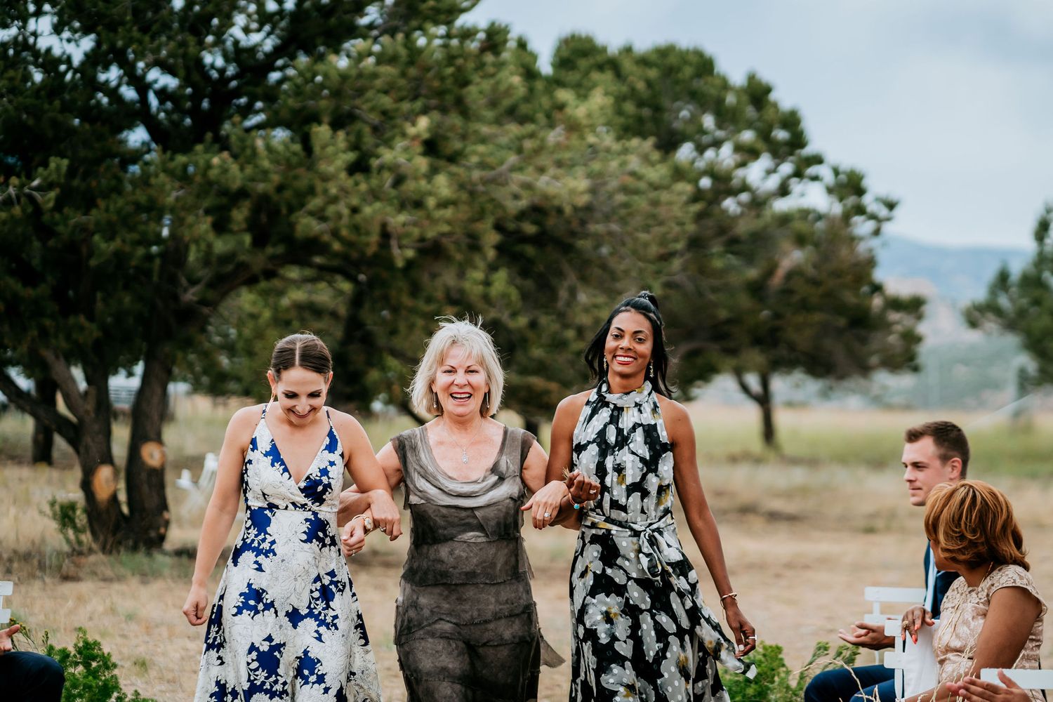 A group walks together outdoors on a grassy hillside wearing formal dresses against a mountain backdrop.