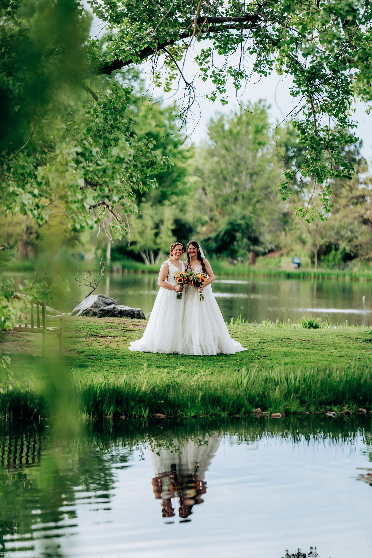 Wedding couple reflected in pond water surrounded by lush green trees.