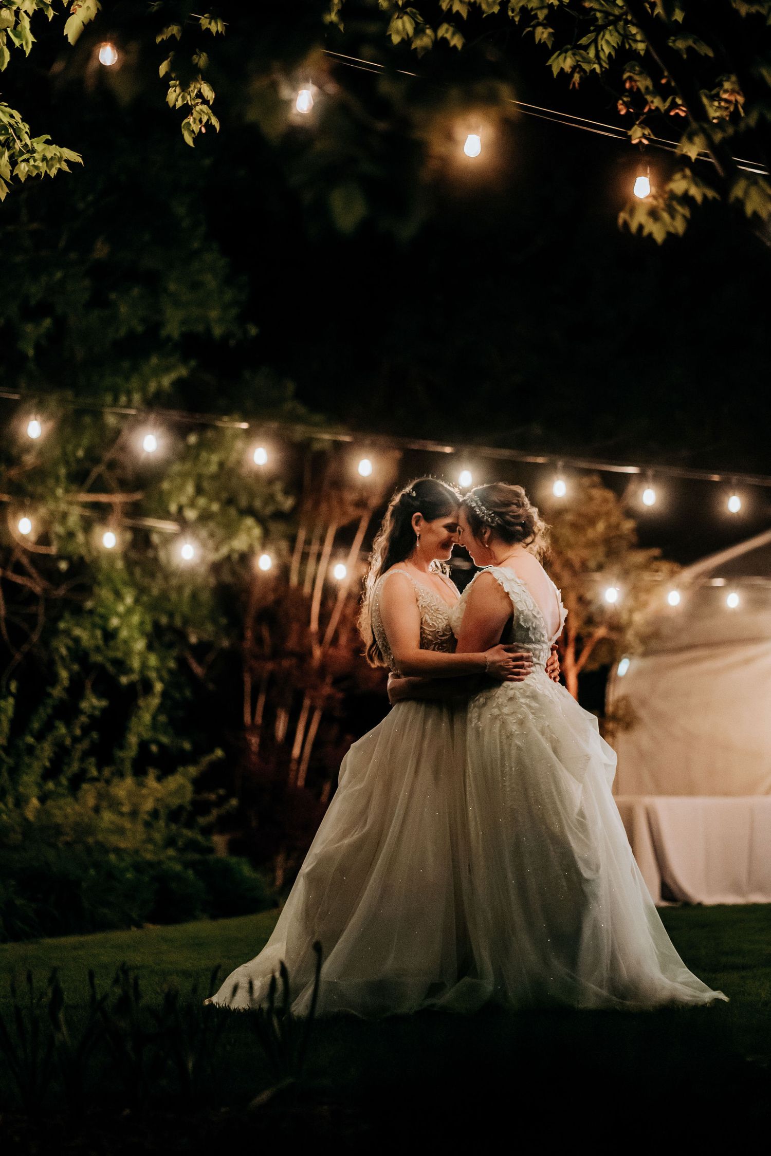 Two brides share an intimate moment under romantic string lights at twilight in a garden setting.