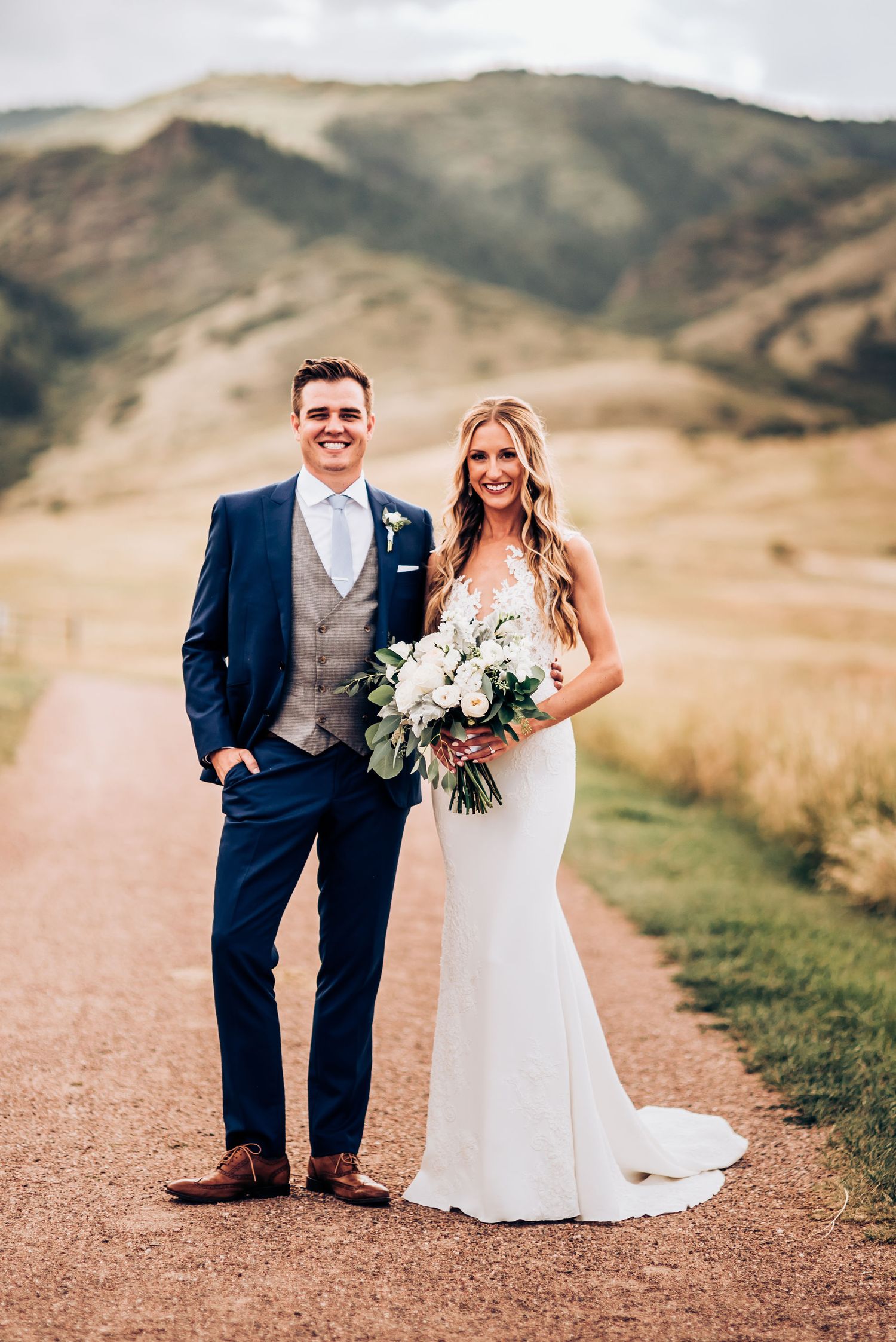 Wedding portrait on mountain trail with bouquet and formal attire against natural backdrop.