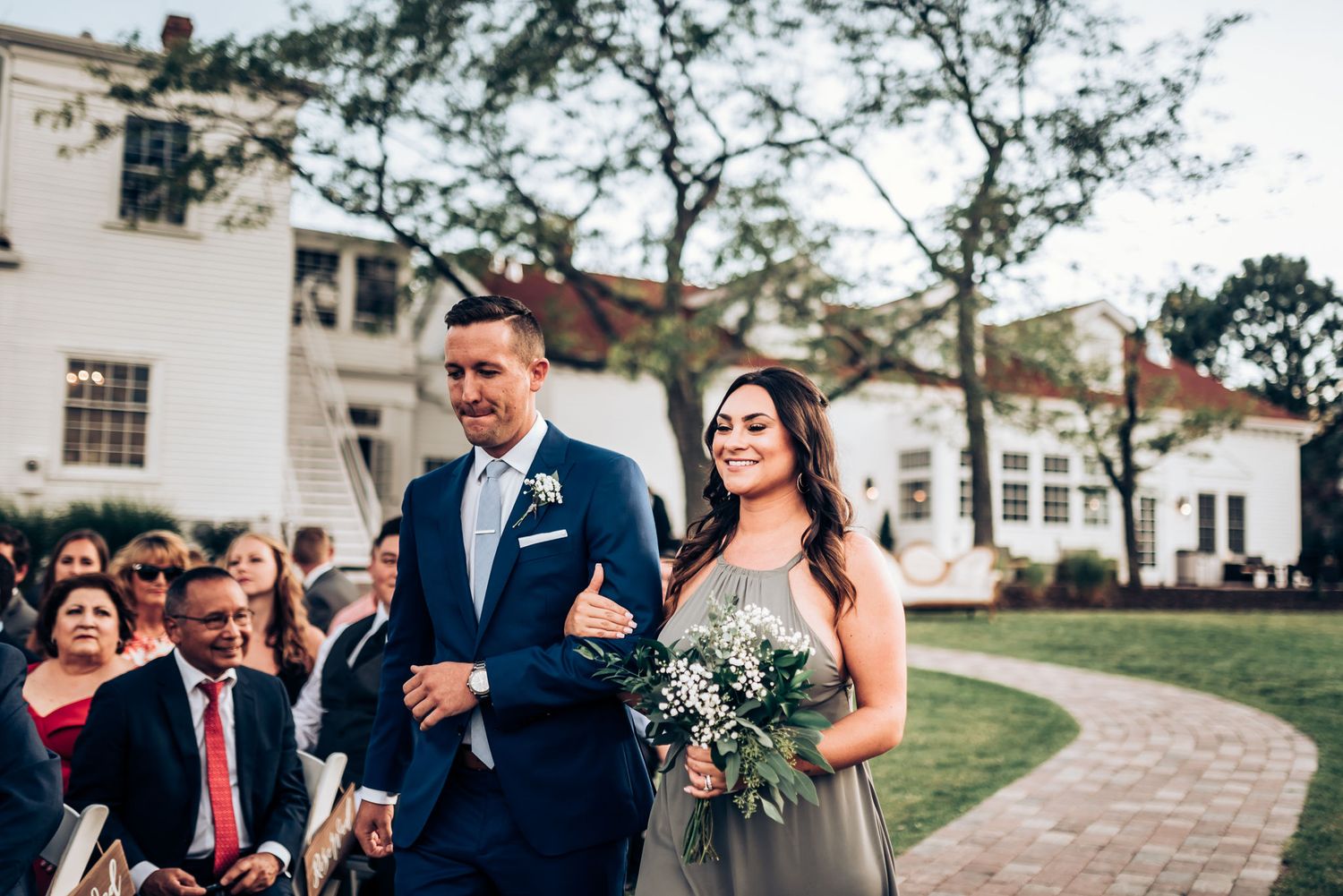 Couple walking processional at outdoor wedding with seated guests and colonial architecture.