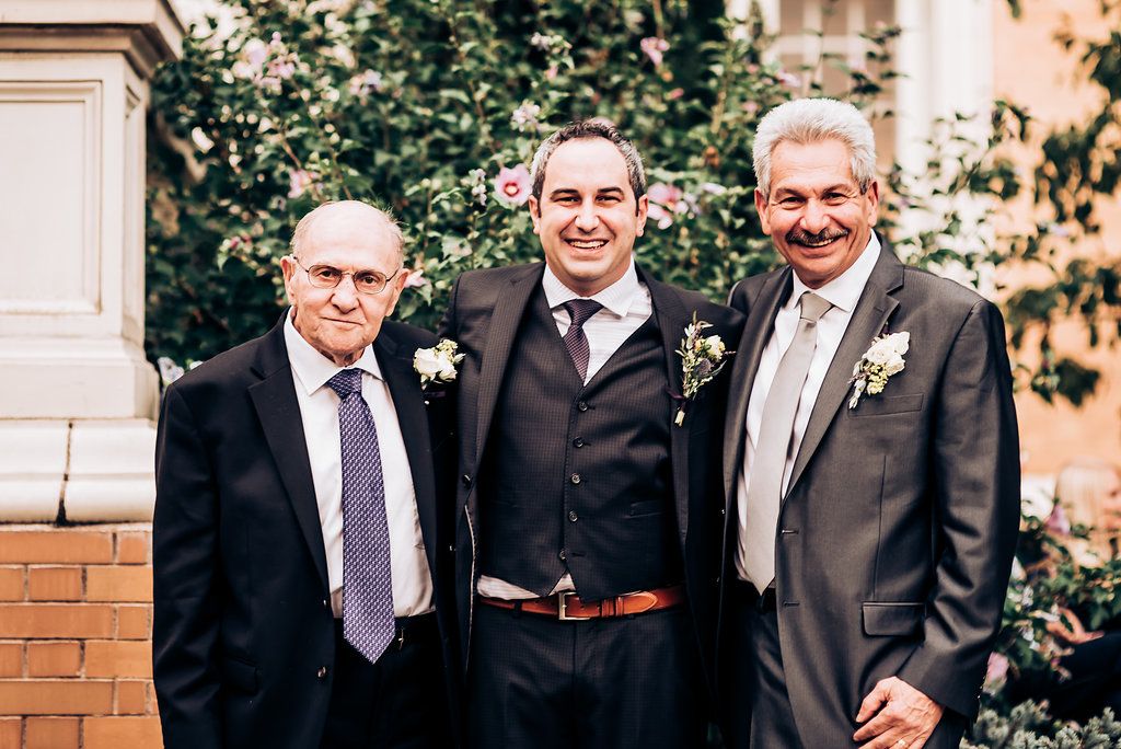 Three formally dressed guests pose together at an outdoor event with brick and greenery.