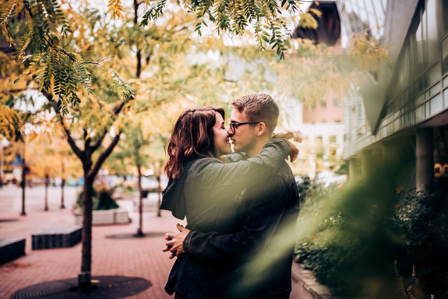 A romantic couple embraces on an autumn city sidewalk with trees in the background.