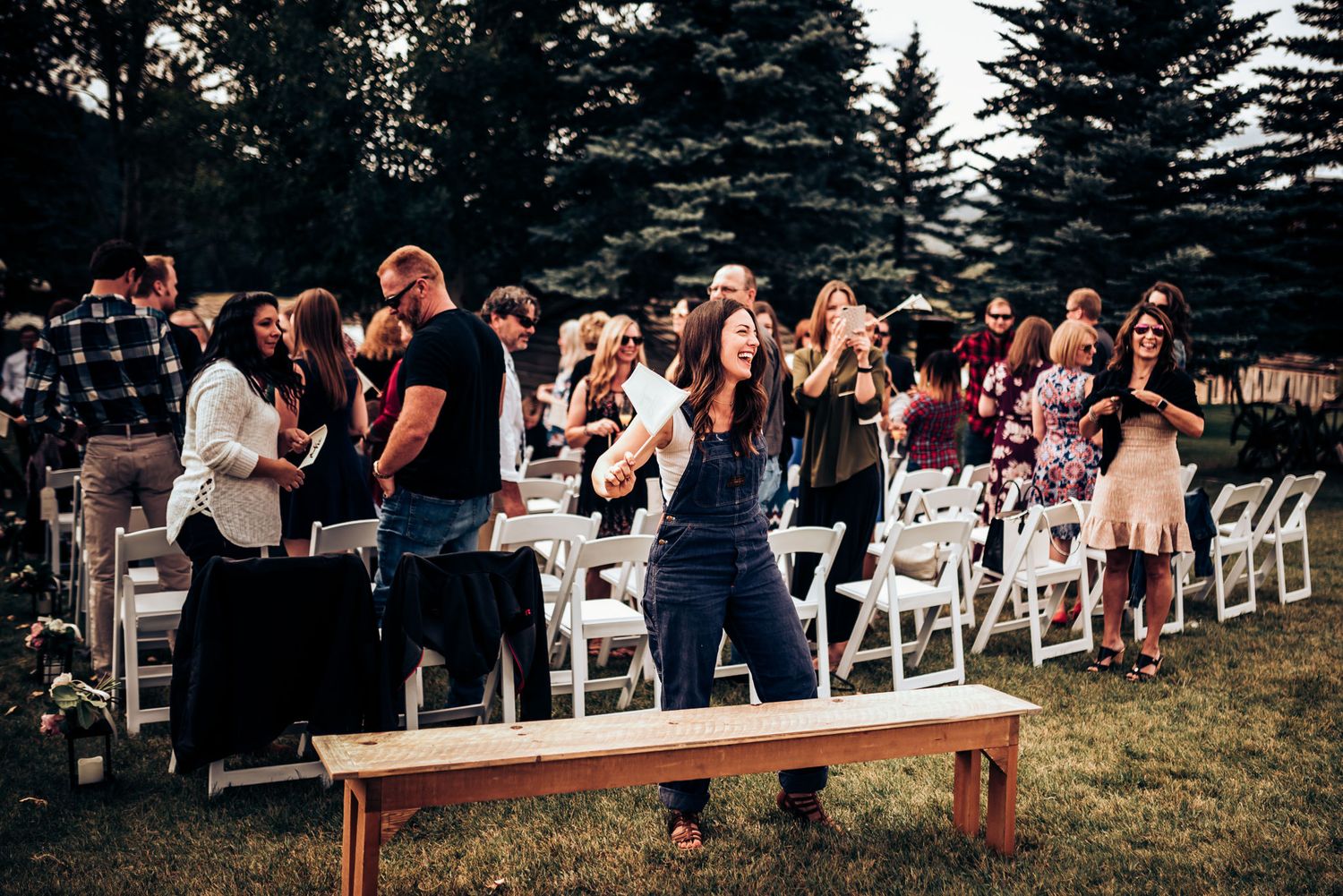 Wedding guests standing and celebrating during an outdoor reception with white chairs and wooden benches.
