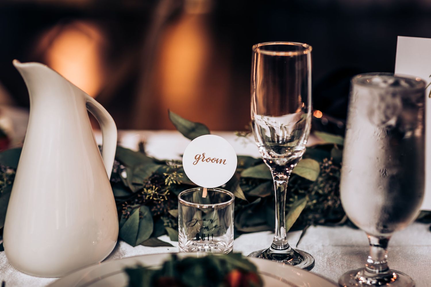 Wedding table detail with white ceramic pitcher, champagne flute and greenery decor.