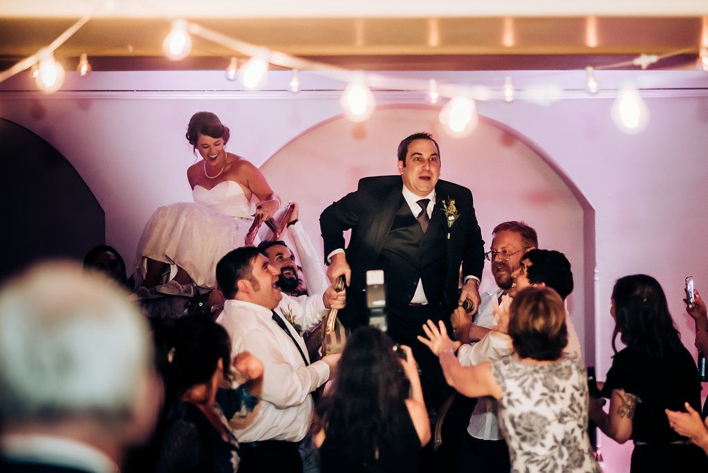 Guests lift and celebrate at an indoor reception under decorative arches and string lights.