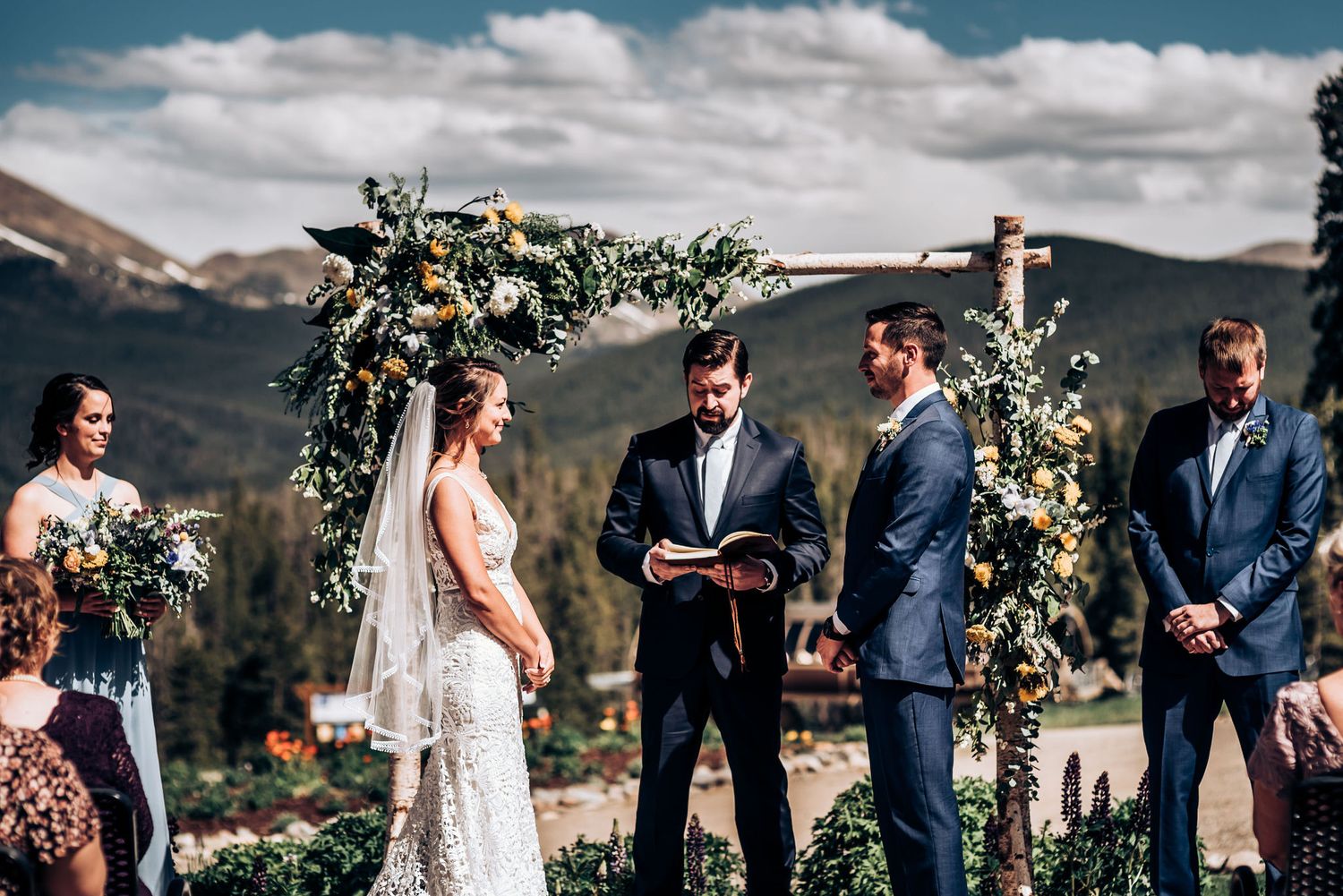 Outdoor wedding ceremony in front of floral arch with mountain landscape in background.