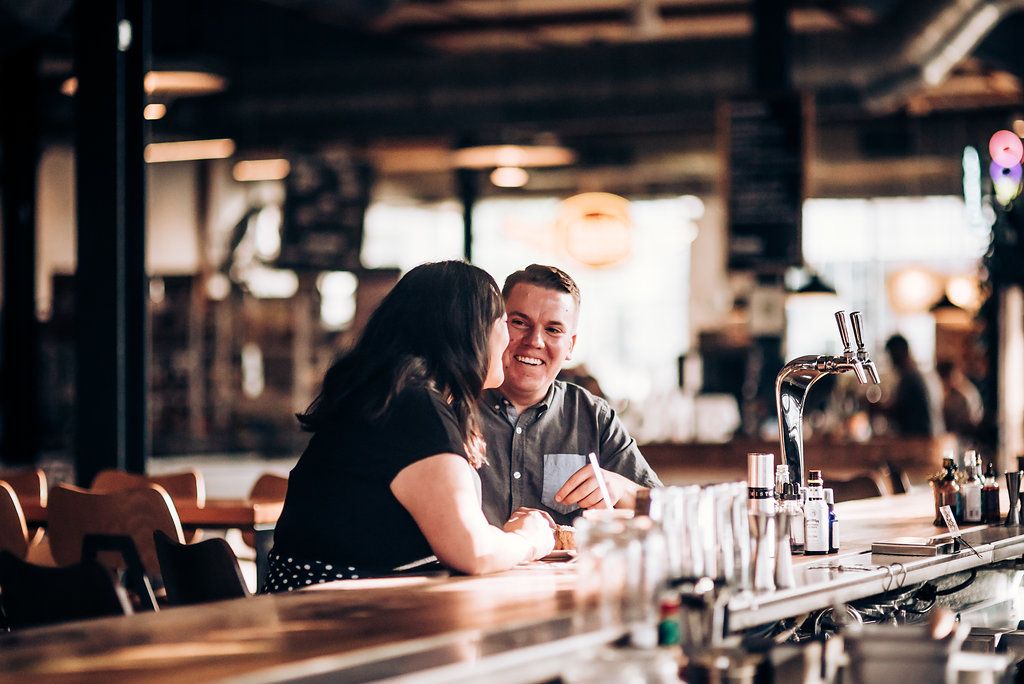 Two people engaged in conversation while seated at a wooden bar counter in a restaurant.
