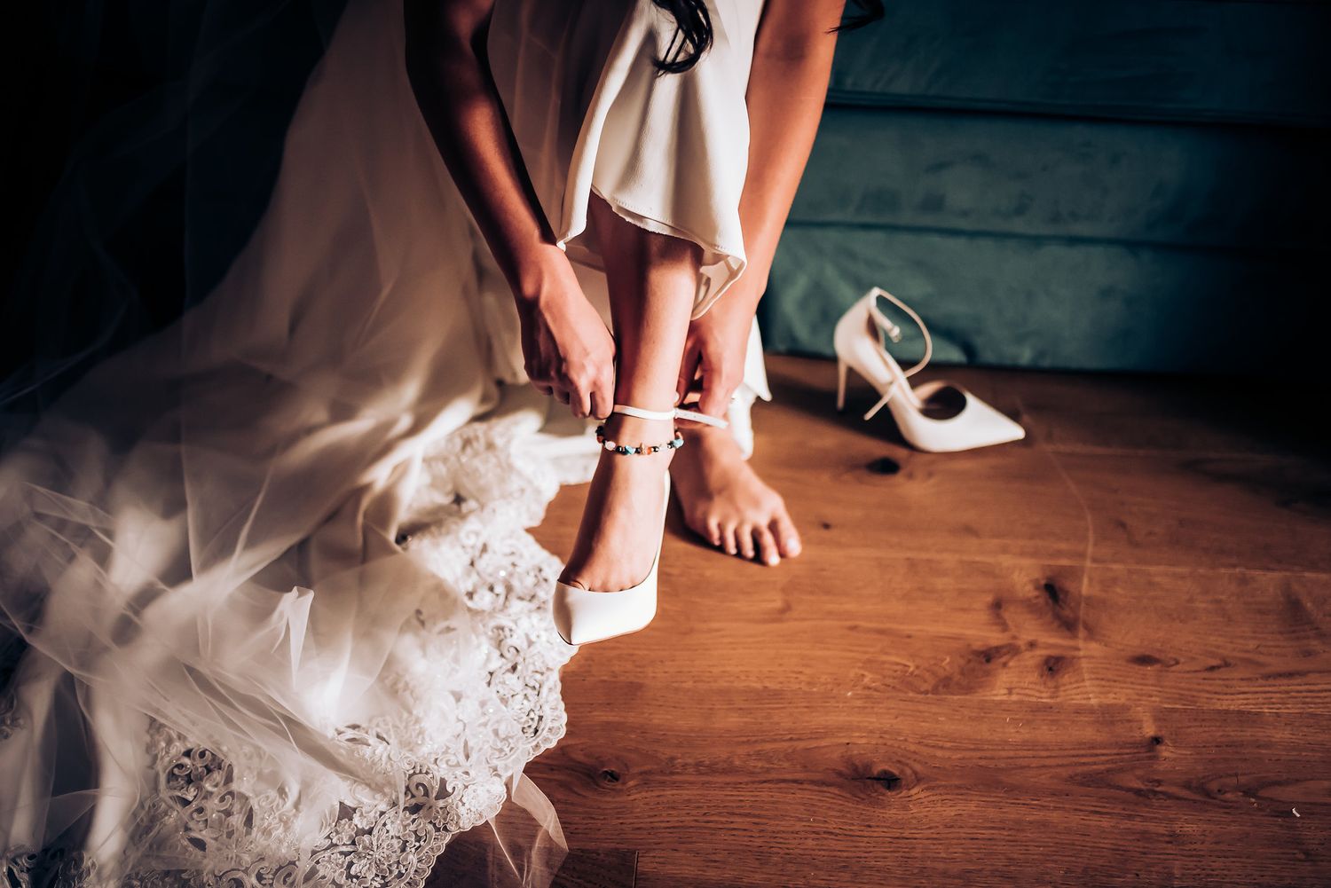 Close-up of elegant white bridal shoes being fastened over lace dress hem on wooden floor.