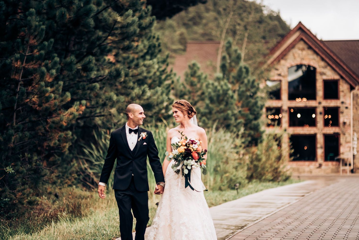 A couple walking together outside a rustic mountain lodge holding bridal bouquet.