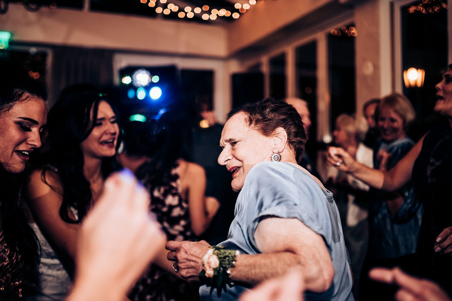 Guests dance enthusiastically on a crowded dance floor under string lights at a celebration.