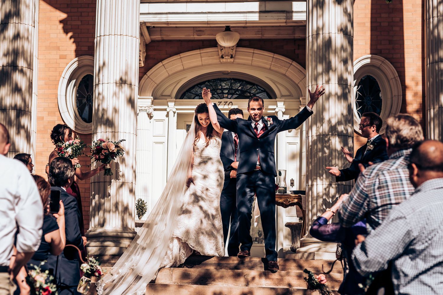 Joyful couple celebrates on historic brick building steps while guests shower them with confetti after wedding ceremony.