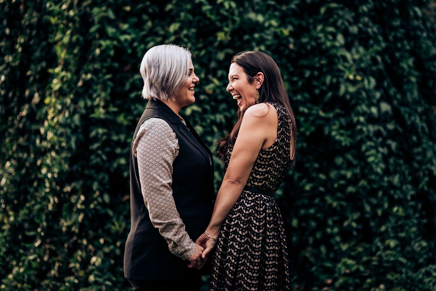 Couple holding hands in front of a lush green hedge wall.