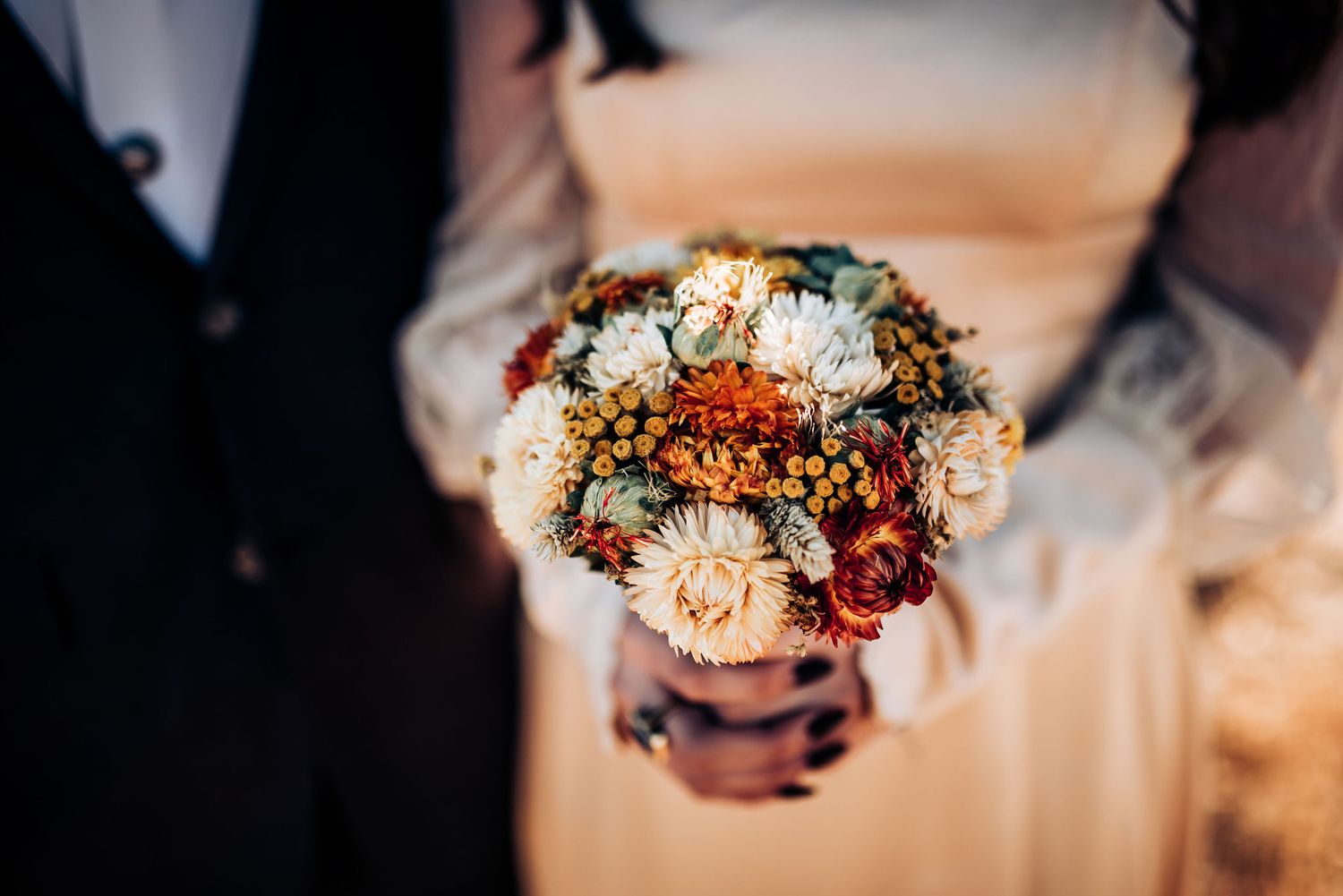 Wedding bouquet with autumn colored flowers and dried elements held against formal attire.