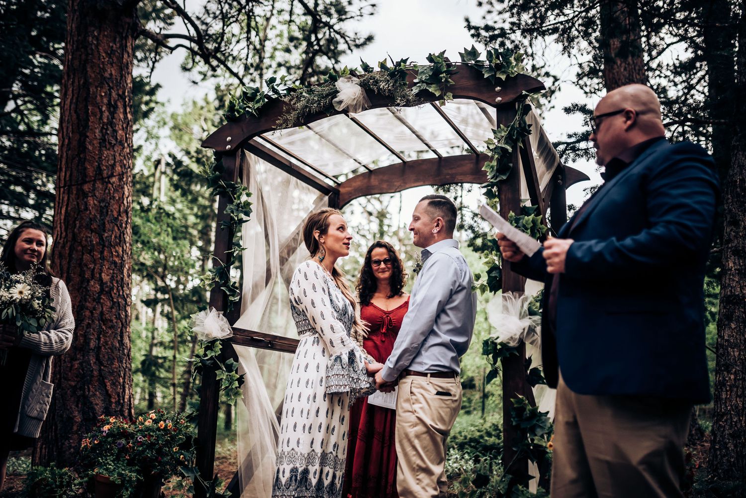 A beautiful woodland wedding ceremony taking place under a decorated wooden arbor.