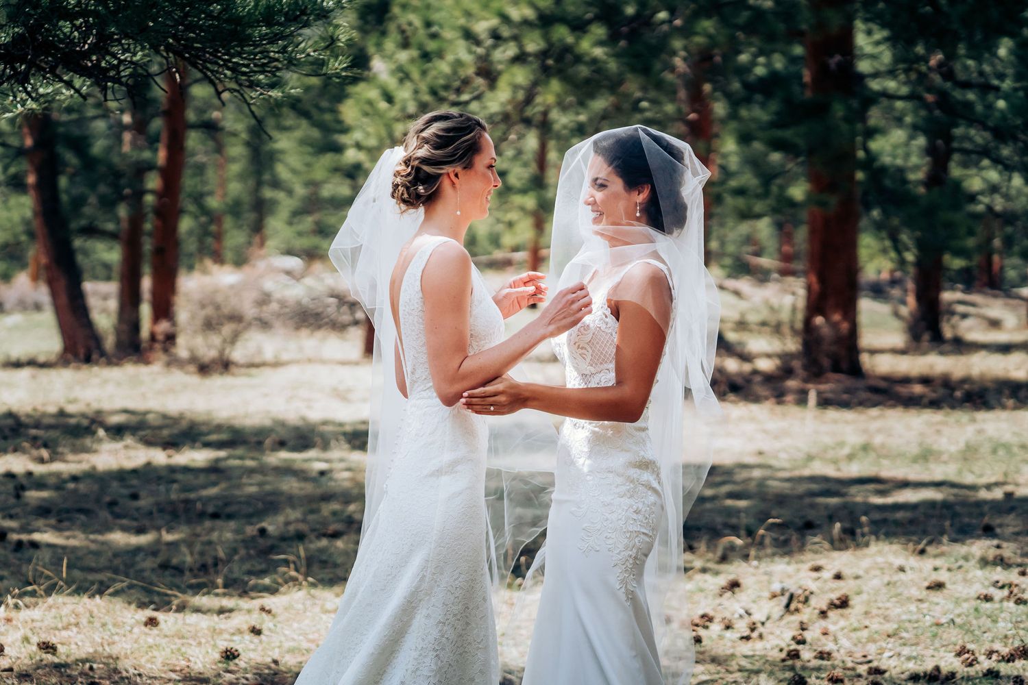 Two people in white wedding dresses hold hands in a sun-dappled forest setting during an outdoor celebration.