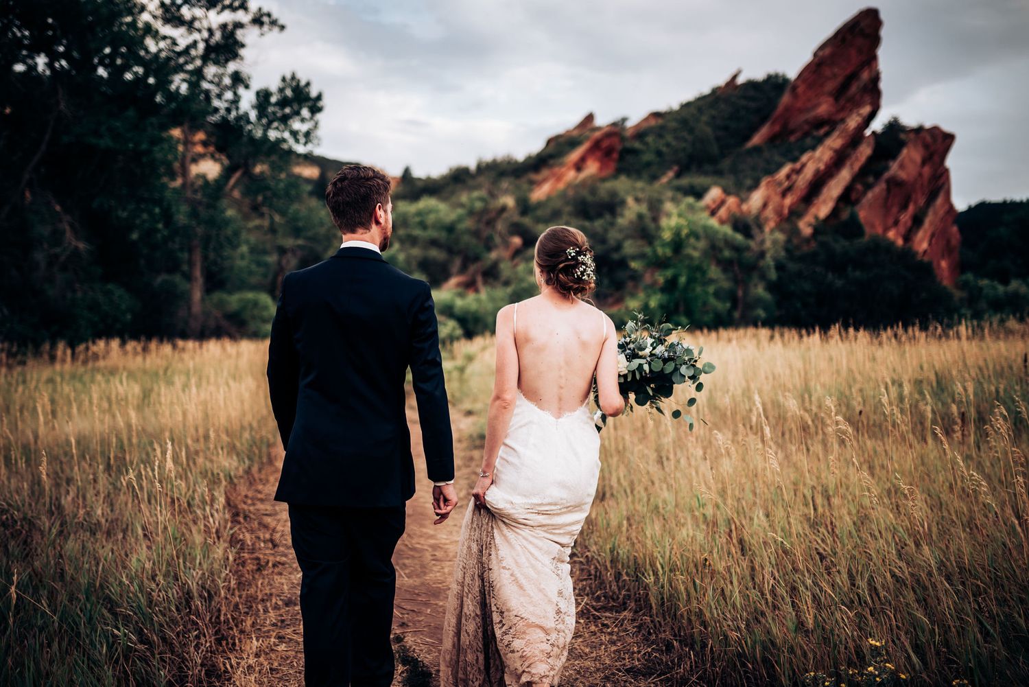 Bride and groom walk through golden field with dramatic red rocks in background.