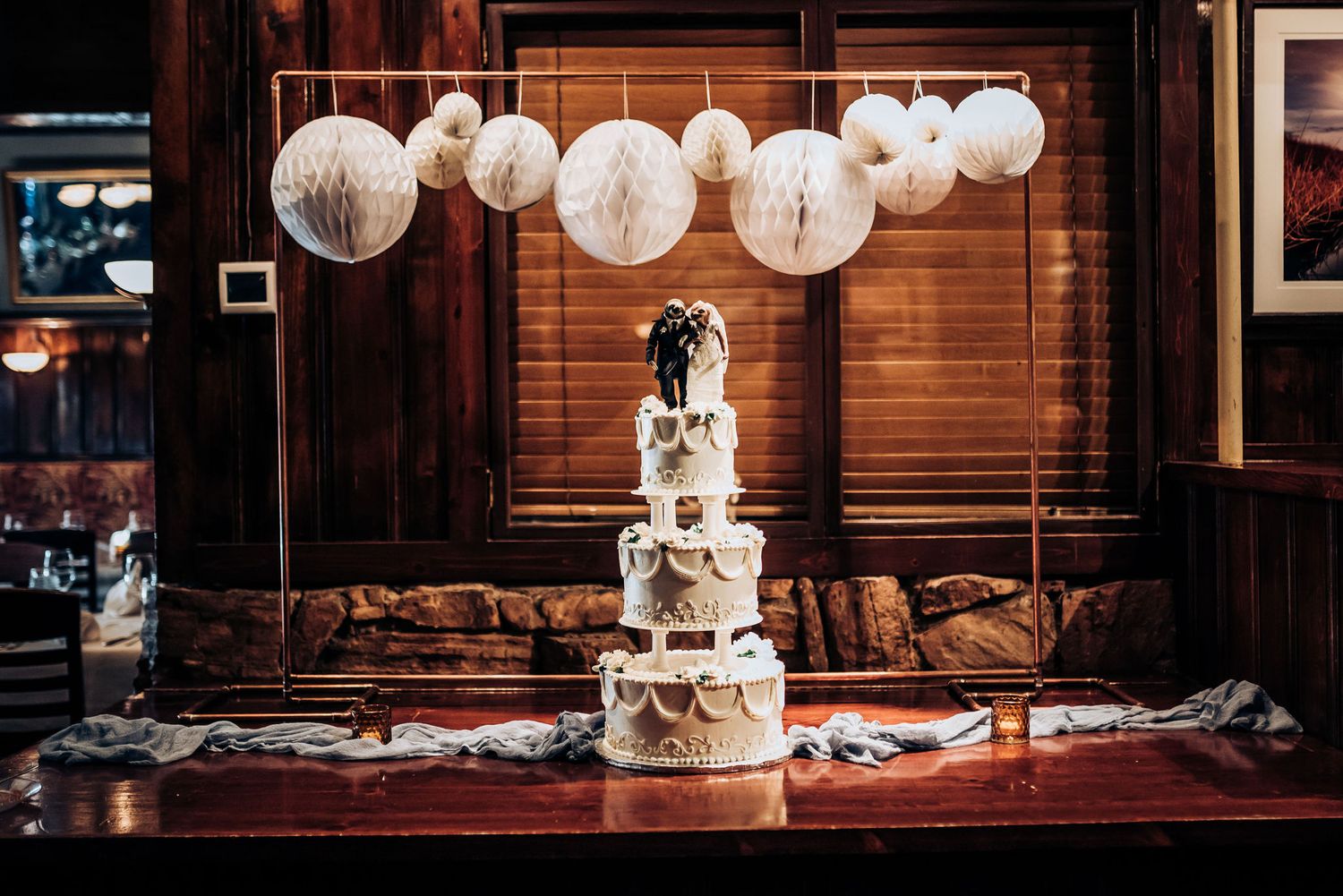 Elegant tiered wedding cake displayed under hanging white paper lanterns in rustic venue.