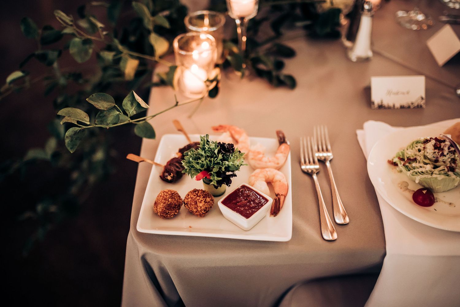 Elegant plated appetizers and silverware on white tablecloth in candlelight.