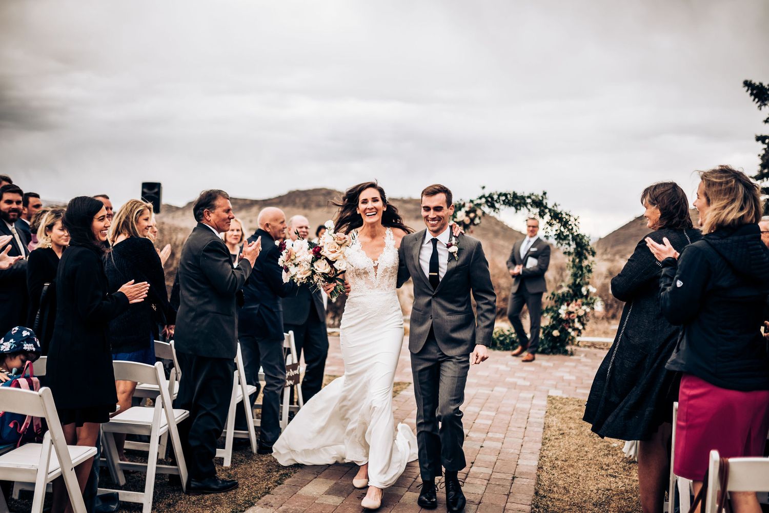 Happy couple exits wedding ceremony while guests in formal attire cheer beneath floral arch.