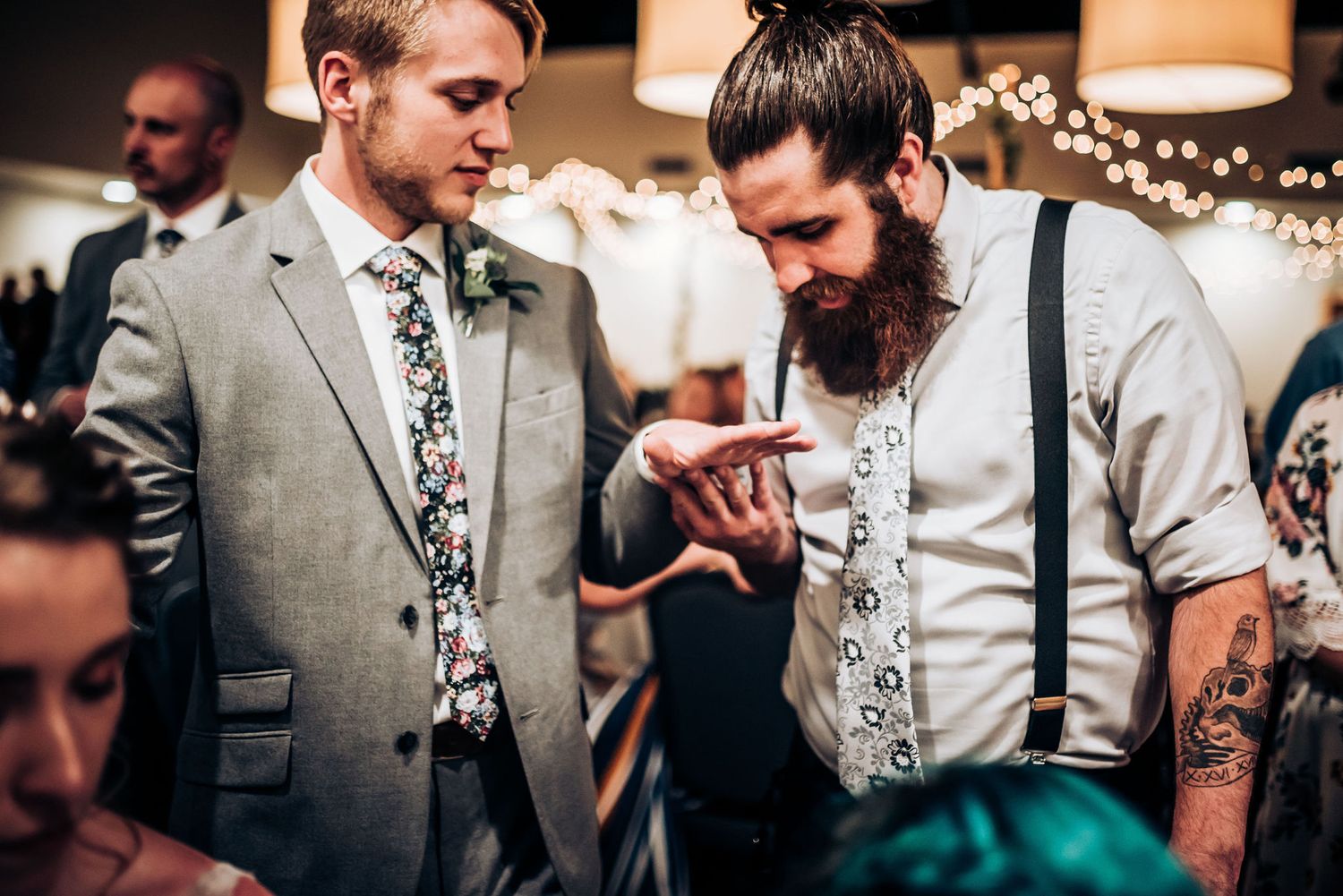 Two guests at a formal event examine something intently under string lights.