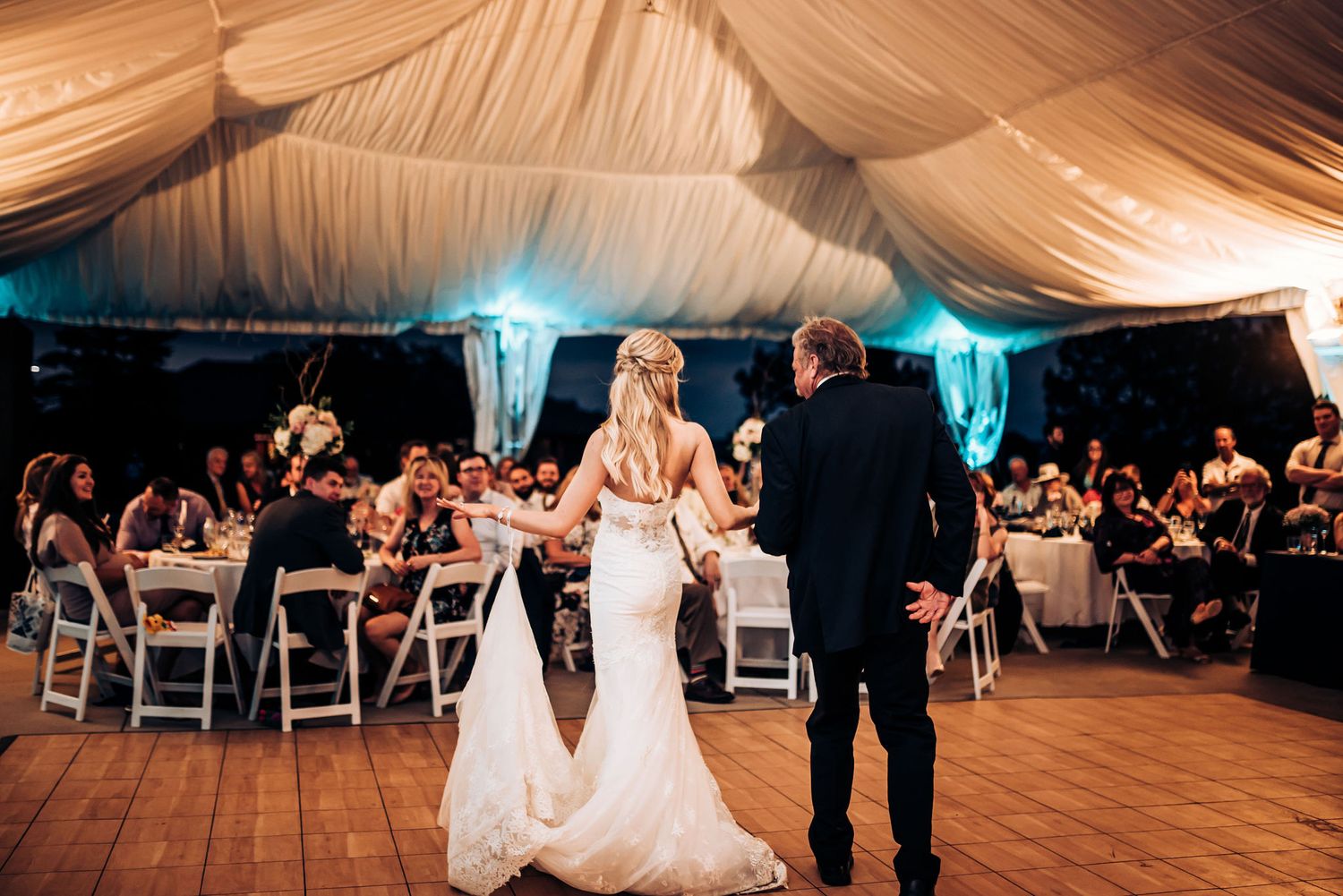 A couple stands before seated wedding guests in an elegantly decorated reception tent with blue accent lighting.