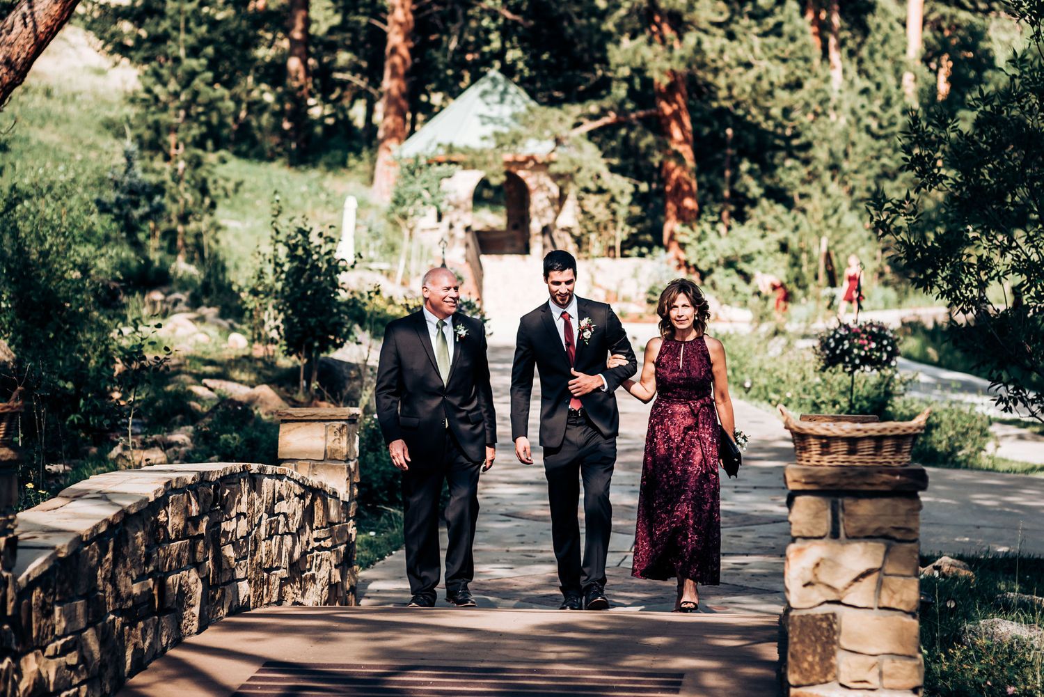 Wedding party members walk along a stone pathway through a scenic garden setting.
