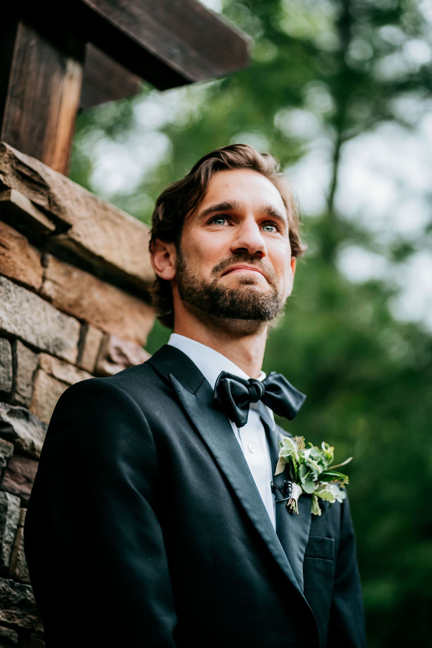 A groom in classic black tuxedo and bow tie poses against a rustic stone wall.