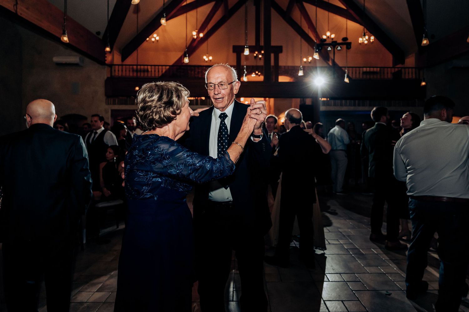 Couples dancing at dimly lit wedding reception with vaulted wood beam ceiling and ambient lighting.
